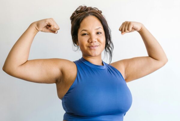 Confident woman in a blue workout outfit flexing her arms and smiling, standing in front of a light background.